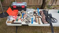 Wide overview of assorted hand tools, toolboxes, tool belt, and work light laid out on a white table outdoors.