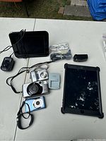 Multiple cameras, Fitbit watch, Kodak Pulse digital frame with OtterBox case, and various chargers and cables displayed on table