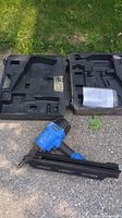 Photo of the blue and black Campbell Hausfeld framing nailer with open black plastic carrying case showing inner compartments and a folded user manual inside the case.