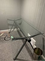 Glass desktop and grey metal frame of the desk seen from a side angle showing the transparent surface and leg design.