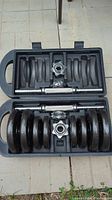 View of the open black plastic case containing two chrome threaded dumbbell bars, multiple black cast iron weight plates, and spin-lock collars all neatly organized inside.