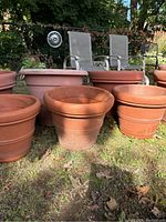 Photo showing seven large plastic planters of different sizes in terra-cotta color placed outdoors on grass with chairs in the background