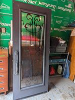 Metal front door with a dark grey finish, iron decorative scrollwork inside a clear glass insert, and a silver handle, photographed in a garage setting with other household items in background.