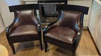 Pair of leather feel armchairs with black wood trim and chocolate brown seats and backs placed on wood floor.