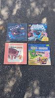Four vinyl album covers displayed on asphalt, featuring Tron sealed album, Science Fiction Gold Star Heroes of Outer Space, Bowmar The Zoo and The Circus, and Pete's Dragon Disney album.
