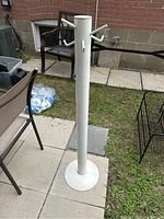 White metal freestanding coat rack shown standing on a patio with background of outdoor furniture and brick wall.