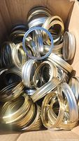 Cardboard box filled with numerous silver metal canning jar screw bands and flat lids.