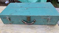 Top view of large rectangular wooden box painted teal with metal latch and worn leather handle, showing exterior condition and closure mechanisms.