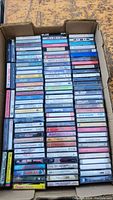 Top-down photo showing about four stacks of cassette tapes neatly arranged in a cardboard box, varied colors and titles visible on side labels.