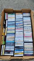 Box containing approximately 60 cassette tapes stacked horizontally and vertically, showing spines with printed titles and artists.
