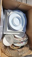 Bird's eye view of the box with assorted kitchenware including white ceramic plates, floral pattern plates with gold trim, a clear glass serving tray, and a white divided dish.