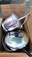 Box containing stacked stainless steel pots and a frying pan with lid, photographed from above showing handles, lids, and reflective surfaces.
