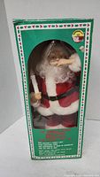 Photo of Santa Claus figurine in original green box with holiday holly pattern and clear window showing figure inside.