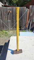 Full view of the sledge hammer showing the length of the yellow handle and metal hammer head on a white surface outdoors.