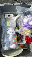 View of storage bin containing assorted collectible items including a white stuffed rabbit plush toy with a red and green scarf in plastic packaging, along with several other smaller items loosely organized inside compartments.