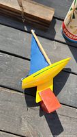 Wooden toy sailboat with blue sail, yellow hull, and red base on a wooden surface.