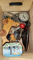 Photo of a wooden toy with round wheels, a dial gauge, a small toy dog, a vintage tin box, and a dark-colored glass or ceramic pedestal object inside a cardboard box.