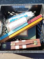 Photo showing black crate with contents: ball of white thread, wooden cribbage board with blue and yellow edges, brown jewelry box.