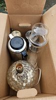 Photo showing vintage textured glass lamp base, Pyrex spaghetti cooker jar with lid, metal coffee pot with black handle and black top, and two copper creamers inside a cardboard box.