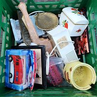 Top down view inside a green milk crate showing a mix of items including hand mirrors, brass decorative pieces, toys, ceramic containers, and a boxed toy fire truck.
