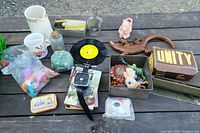 Photo showing various items on wooden table including toys in plastic bags, ceramic mugs, glass bottle, vinyl record, wooden door handle with lock, small tin, rock specimen, and box labeled 'UNITY'.