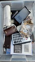 Wide view of plastic bin containing EL calculator, black electronic device, brown leather pouches, and decorative hardware pieces in plastic.