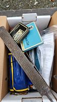 Box of assorted hand tools including a metal rasp, blue pouch, tin containers, and other tools.