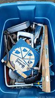 Blue bin filled with assorted metal hand tools including a Woodward's Hardware circular saw blade, wooden and metal handled saws, measuring tools, wrenches, and other hand tools.