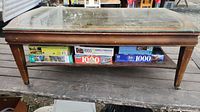 Photo of weathered wooden table with glass top and decorative gold border. Lower shelf showing multiple puzzle boxes.