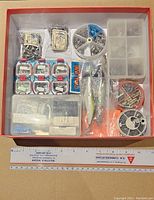 Top view showing assortment of fishing tackle items including hooks, weights in plastic containers, small lures, and multiple small packages inside a red bordered box.