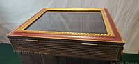 Closed view of the storage box showing red and gold finished wood lid with glass top, gold rope accent bordering the glass pane.