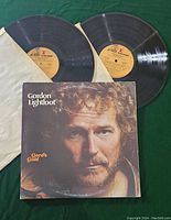 Two black vinyl LP records partially removed from protective paper sleeves, with yellow center labels showing album and track information. Gord's Gold album cover featuring Gordon Lightfoot's face in close-up is visible.