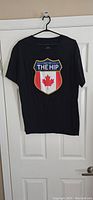Black T-shirt hanging on a hanger showing the front with a colorful shield logo with a red maple leaf and text.