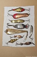 Collection of nine vintage fishing spoons arranged on white cloth showing variety in shapes, sizes, finishes and hooks.