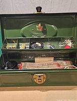 Green metal fishing tackle box open showing tray with various vintage fishing lures, fishing line spool, bobbers, and other fishing accessories inside.