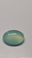 Oval cabochon shaped chalcedony gemstone with arctic teal blue color on a neutral background, showing polished surface and clarity.