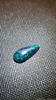 Teardrop-shaped polished black opal with visible flashes of green and blue colors on textured black background.