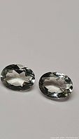 Pair of faceted oval cut teal colored green amethyst gemstones on a white background showing facets and transparency.