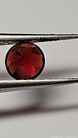 Close-up image of a round cut deep red garnet gemstone held in tweezers, showing the gem face on with visible facets and internal clarity.