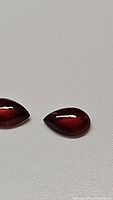 Close-up photo of two pear-shaped cherry red garnet cabochon gemstones on a plain background, showing smooth polished surfaces and matching shapes.