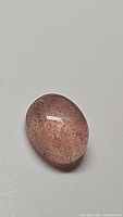 Close-up photo of oval cabochon cut natural pink strawberry quartz showing internal mineral inclusions and soft pink color on white background.
