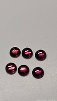 Six natural round cabochon Rhodolite garnet stones arranged on a neutral surface showing pink-purple hues with glossy finish and good internal clarity.