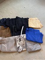 Nine pairs of assorted women's pants displayed on carpet, showing colors and patterns including solid black, blue, tan, cream, gray, stripes, and florals.