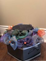 Black woven straw hat topped with multicolored organza flower accents on hexagonal box