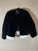 Front view of black faux fur jacket with white hem trim on wooden hanger