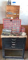View of black Craftsman roller cabinet with three drawers and open bottom, small wooden shelf unit above, plastic storage drawer organizer, and various tools on top.