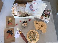 Full lot view showing crocheted doilies, needlepoint squares, two small dolls, porcelain doll parts, wooden puzzles, and illustrations