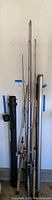 Photo showing multiple fishing rods of various lengths leaned against a wall with two carrying tubes, one metal and one black plastic.