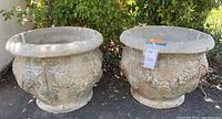 Two large round concrete planters with grapevine motif shown outdoors on pavement with greenery background. Both planters exhibit weathered surface patina in celadon, taupe, and light terra-cotta colors.