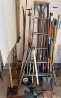 Full view of lot showing wooden ladder, hand tools including shovels, spades, rakes, pitchforks, axe and Black & Decker hedge trimmer on floor.